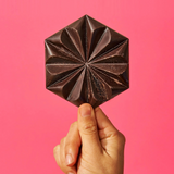 A hand holding a handcrafted, hexagonal single-origin chocolate tablet against a vibrant pink background. The tablet features a decorative floral-like pattern, showcasing artisanal Mexican chocolate craftsmanship.