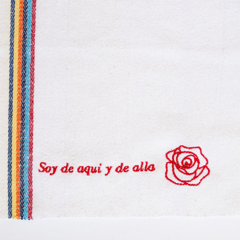 A close up of a white recycled cotton cleaning cloth reading "Soy de aqui y de alla" in red with a red rose embroidered next to it