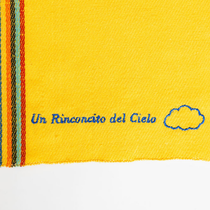 A close up of a yellow recycled cotton cleaning cloth with blue embroidery reading "Un Rinconcito del cielo" and a blue cloud