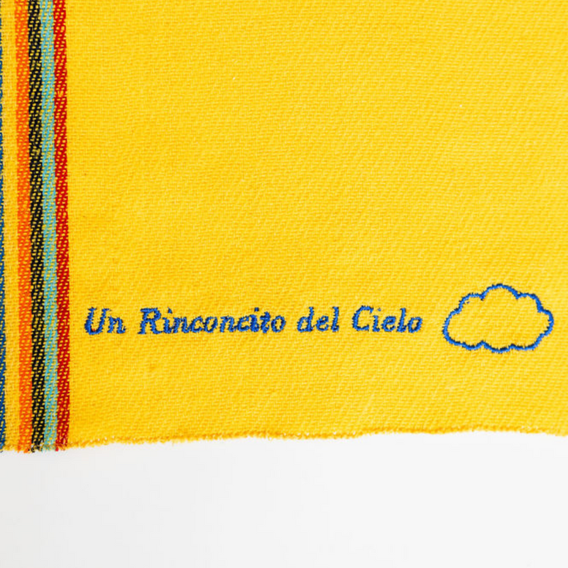 A close up of a yellow recycled cotton cleaning cloth known as Jerga with blue embroidery reading "Un Rinconcito del cielo" and a blue cloud