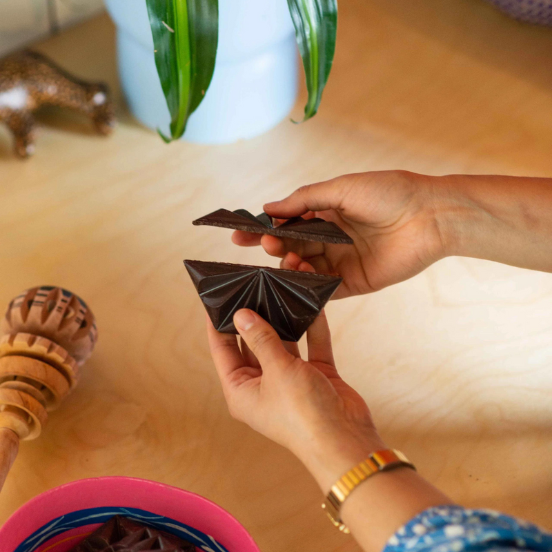 Hand breaking apart artisanal Mexican chocolate tablet, used for traditional drinking chocolate, with a molinillo whisk nearby. Made with single-origin Mexican cacao using heritage techniques.