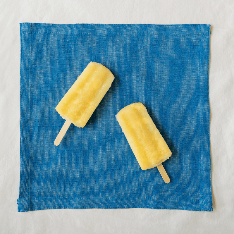 A teal cotton woven napkin laying flat with 2 yellow paletas sitting on top of it