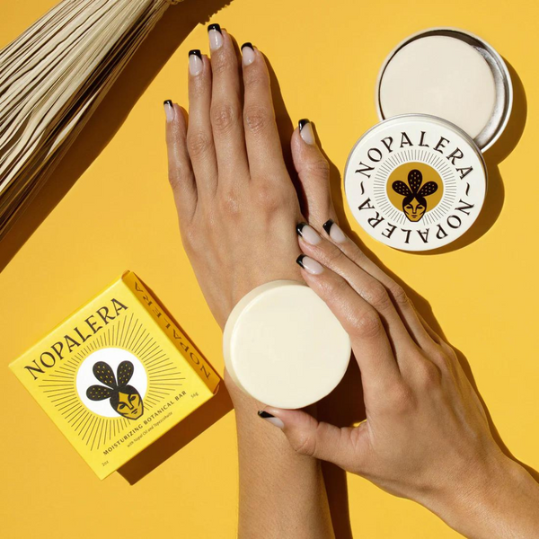 Nopalera's Original Moisturizing Botanical Lotion Bar  being applied on a hand as lotion on a yellow background