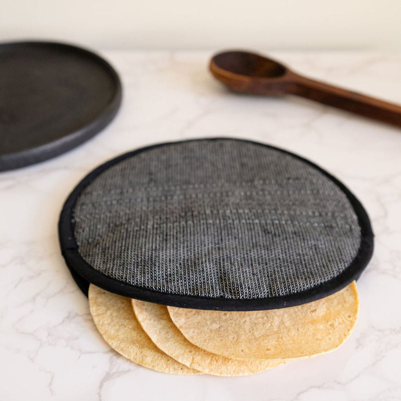 A Black Handwoven Cotton Tortilla Warmer sits elegantly on a table, showcasing its natural, dark tones and intricate handwoven texture. Made from 100% cotton, this eco-friendly warmer adds a rustic, artisan touch to any meal while keeping tortillas warm and fresh.