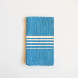 A folded Casa Oaxaca Cotton Cloth Napkin in a striking Blue Azure shade. The napkin features a minimalist design with five evenly spaced white lines of various thickness running horizontally across its surface, creating a subtle yet elegant contrast against the deep blue. The napkin's texture showcases its artisanal craftsmanship, made from 100% Mexican cotton, offering a blend of durability and softness. 