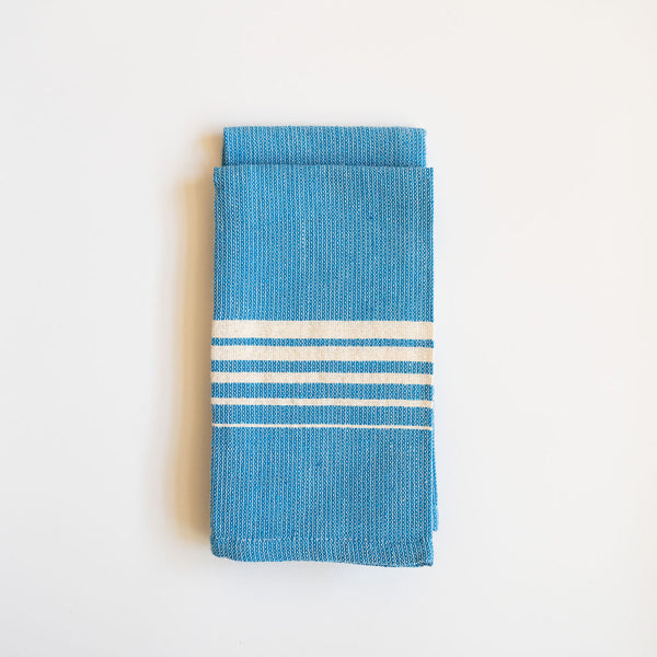 Set of two folded Casa Oaxaca Cotton Cloth Napkin in a striking Blue Azure shade. The napkin features a minimalist design with five evenly spaced white lines of various thickness running horizontally across its surface, creating a subtle yet elegant contrast against the deep blue. The napkin's texture showcases its artisanal craftsmanship, made from 100% Mexican cotton, offering a blend of durability and softness. 