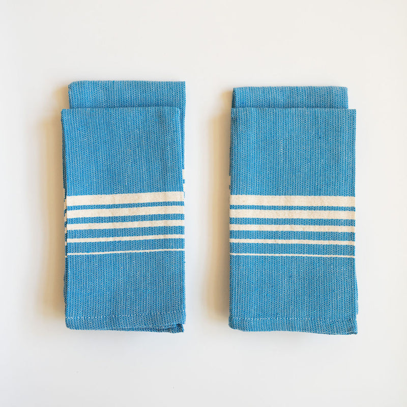 Set of four folded Casa Oaxaca Cotton Cloth Napkin in a striking Blue Azure shade. The napkin features a minimalist design with five evenly spaced white lines of various thickness running horizontally across its surface, creating a subtle yet elegant contrast against the deep blue. The napkin's texture showcases its artisanal craftsmanship, made from 100% Mexican cotton, offering a blend of durability and softness. 