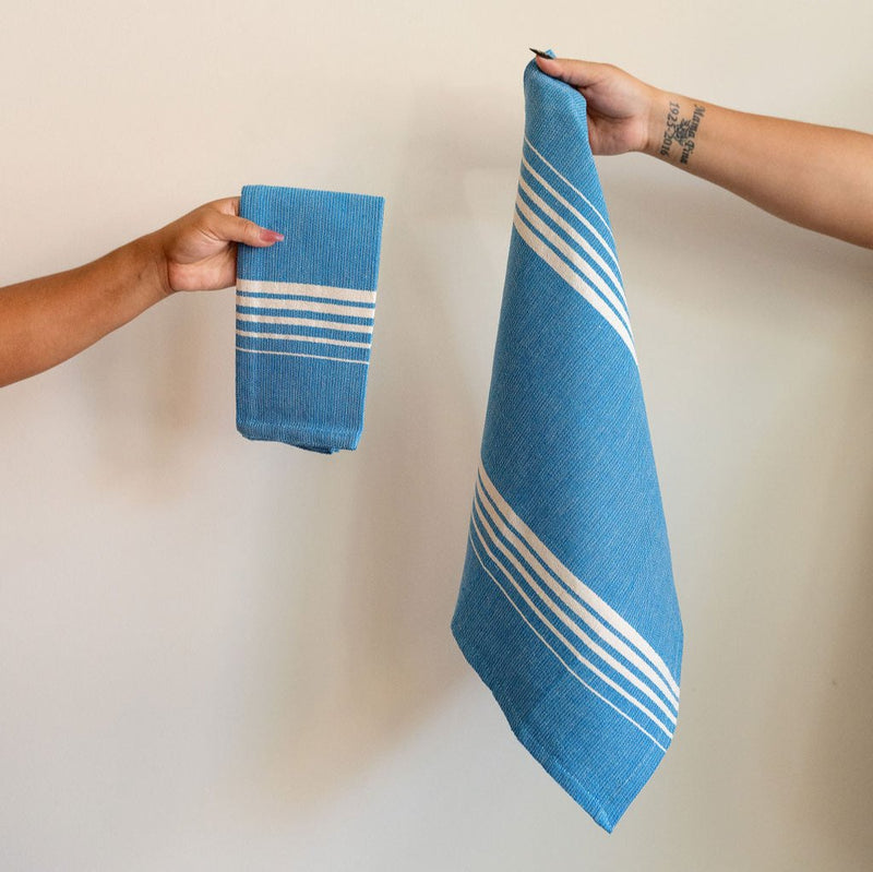 Set of two folded Casa Oaxaca Cotton Cloth Napkin in a striking Blue Azure shade. The napkin features a minimalist design with five evenly spaced white lines of various thickness running horizontally across its surface, creating a subtle yet elegant contrast against the deep blue. The napkin's texture showcases its artisanal craftsmanship, made from 100% Mexican cotton, offering a blend of durability and softness. 