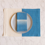 A Blue Azure Casa Oaxaca Cotton Cloth Napkin, featuring five delicate white lines, rests elegantly on a sleek gray plate, perfectly paired with a matching woven placemat. This handwoven napkin adds a pop of vibrant blue to any sustainable table setting, ideal for eco-conscious homes.