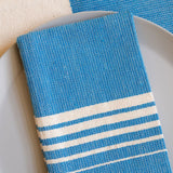 A Blue Azure Casa Oaxaca Cotton Cloth Napkin, featuring five delicate white lines, rests elegantly on a sleek gray plate, perfectly paired with a matching woven placemat. This handwoven napkin adds a pop of vibrant blue to any sustainable table setting, ideal for eco-conscious homes.