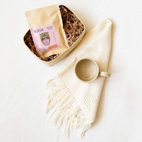 A handwoven palm basket filled with crinkled paper and a packet of Hijita Mexican cacao sits on a light surface beside a cream-colored, fringed cotton hand towel with subtle woven lines. A matching ceramic mug rests on top of the towel, creating a warm, artisanal gift set.