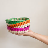 Mexican colorful palm baskets with natural bottom and colored rims: pink (Rosa Mexicano), Orange (Apricot), and green (Emerald Green) stacked on top of each other held by a hand