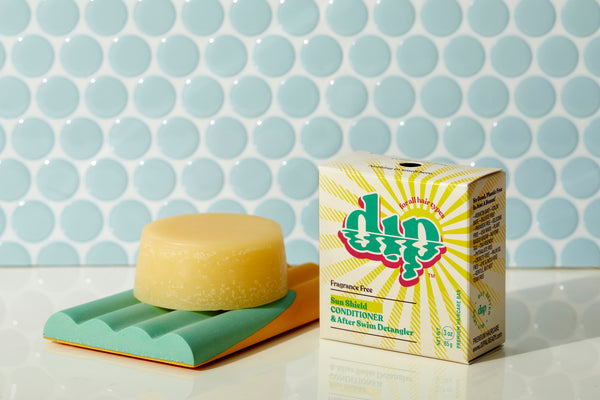 A fragrance-free sun shield dip conditioner bar in its eco-friendly box, with a second bar sitting on a smooth surface beside it. The packaging has a clean, minimalist design with the text "Fragrance Free Sun Shield Dip Conditioner Bar" clearly visible. The bars have a solid, creamy texture, perfect for protecting and conditioning hair without added scents.