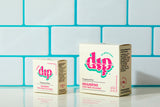 A mini fragrance-free dip shampoo bar sits next to a full-sized version, both in their eco-friendly boxes. The packaging showcases clean, simple design elements with "Fragrance Free Shampoo Color Safe" clearly labeled. The bars, with their smooth and solid textures, highlight their compact, travel-friendly, and full-sized options.