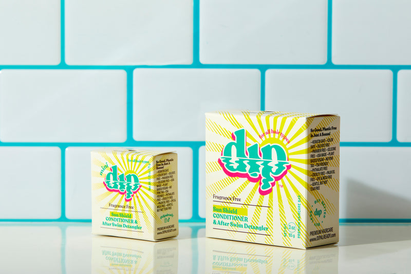 A mini fragrance-free Sun Shield dip conditioner bar sits next to a full-sized version, both in their eco-friendly boxes. The packaging showcases clean, simple design elements with "Fragrance Free Sun Shield Dip Conditioner Bar" clearly labeled. The bars, with their smooth and solid textures, highlight their compact, travel-friendly, and full-sized options for effective sun protection and conditioning.