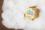 A coconut and almond dip shampoo bar in its box, surrounded by sudsy soap bubbles. The shampoo bar is eco-friendly and packaged in minimal, sustainable materials, highlighting its natural ingredients and low-waste benefits.