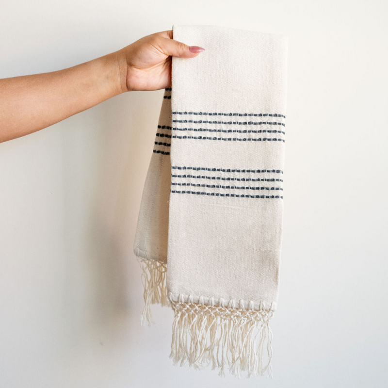 Cotton Woven Kitchen Hand Towel in natural with slate grey stripes and 100% cotton fringe held by a hand against a wall