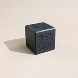 A dark charcoal gray vegan organic cube-shaped bar of soap set against a warm beige background with a soft shadow.