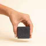 A hand holding a dark charcoal gray vegan organic cube-shaped bar of soap set against a warm beige background with a soft shadow.