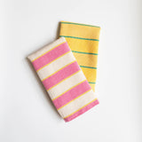 Two folded cotton napkins arranged on a white background. The top napkin features cream/off-white fabric with wide pink stripes and thin yellow accent lines. The bottom napkin displays a bright yellow fabric with thin green horizontal stripes. Both textiles showcase traditional Mexican cotton weaving techniques with vibrant colors and striped patterns