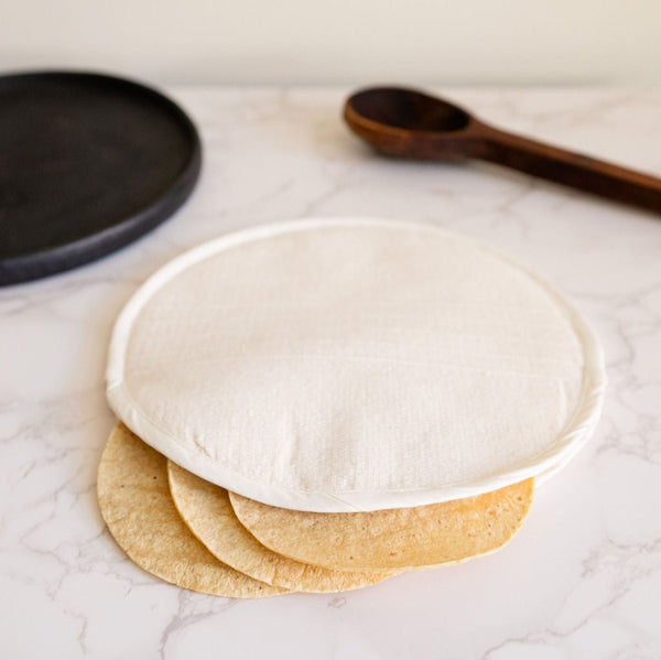 A Natural Creme Handwoven Cotton Tortilla Warmer sits elegantly on a table, showcasing its soft, neutral tone and artisanal texture. Made from 100% cotton, this eco-friendly warmer adds a traditional yet minimalist touch to any kitchen or dining space. The Warmer has tortillas inside and you can see a plate and wood spoon in the background.