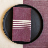 A Purple Jacaranda Casa Oaxaca Cotton Cloth Napkin, featuring five delicate white lines, sits elegantly on a black plate, perfectly paired with a matching woven placemat. This vibrant, handcrafted napkin adds a touch of artisanal Mexican charm to any eco-friendly table setting.