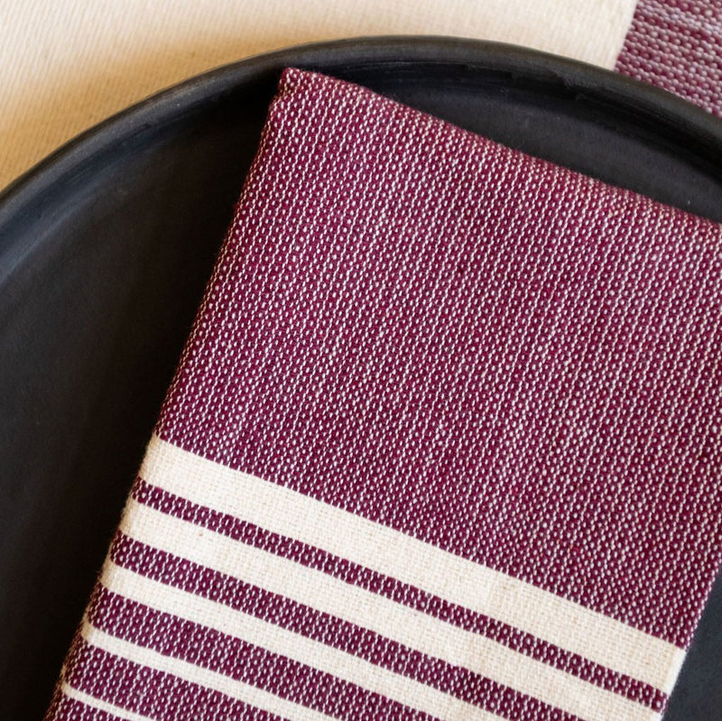 A Purple Jacaranda Casa Oaxaca Cotton Cloth Napkin, featuring five delicate white lines, sits elegantly on a black plate, perfectly paired with a matching woven placemat. This vibrant, handcrafted napkin adds a touch of artisanal Mexican charm to any eco-friendly table setting.