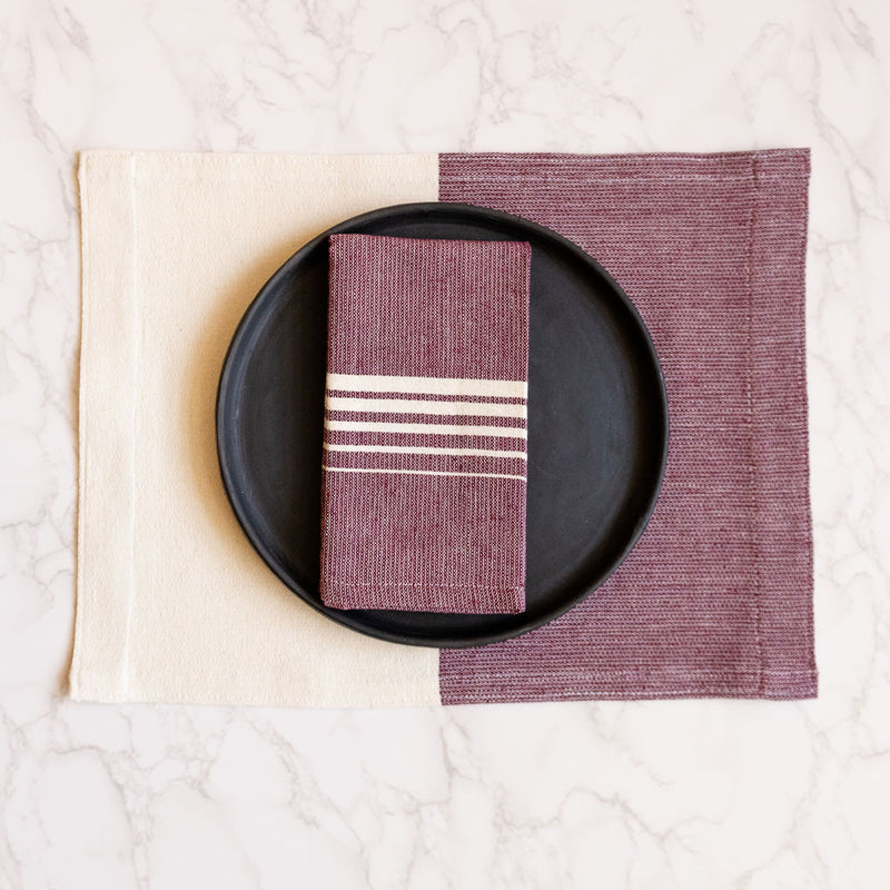 A vibrant Purple Jacaranda Casa Oaxaca Woven Cotton Placemat, handwoven in Oaxaca, serves as the backdrop for a sleek black plate, topped with a matching Jacaranda napkin. This artisanal Mexican table setting adds a bold pop of color, perfect for eco-conscious homes seeking unique, handcrafted decor.