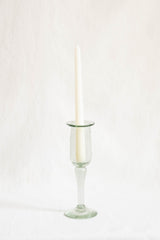 A taper candle standing in a clear glass candlestick holder with a flared base, photographed against a soft off-white fabric background.