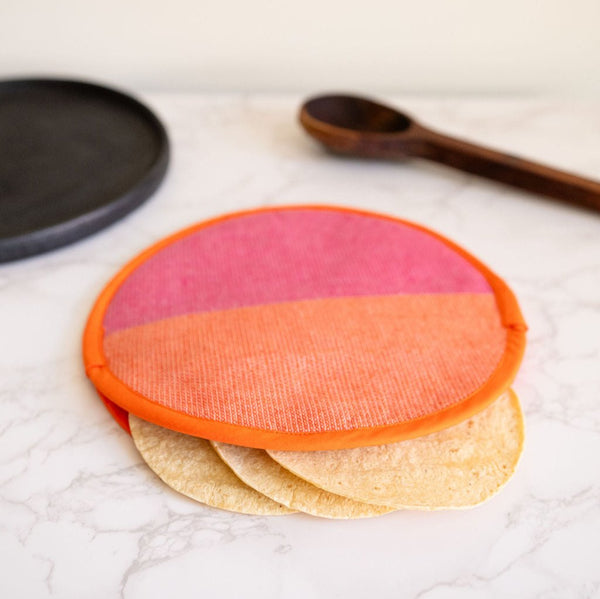 A vibrant Rosa Mexicano pink and orange color block handwoven cotton tortilla warmer, perfect for keeping tortillas warm while adding a pop of color to your table. This eco-friendly, artisan-made kitchen accessory is crafted from sustainable materials, reflecting Mexican cultural heritage and zero-waste values. With tortillias inside and a black plate and wood spoon in the background.