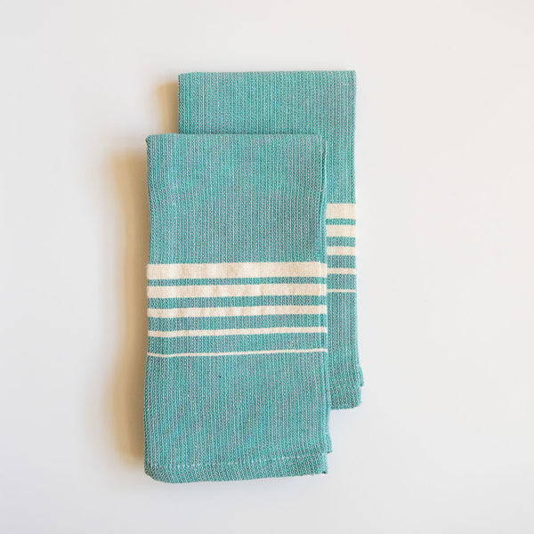 A vibrant Set of Two Teal Casa Oaxaca Woven Cotton Napkin, crafted from 100% Mexican cotton, adds a pop of color to any table setting with its artisanal design and sustainable, eco-friendly material. Perfect for elevating your dining experience, this napkin is handwoven in Oaxaca and designed to complement a modern, conscious lifestyle.