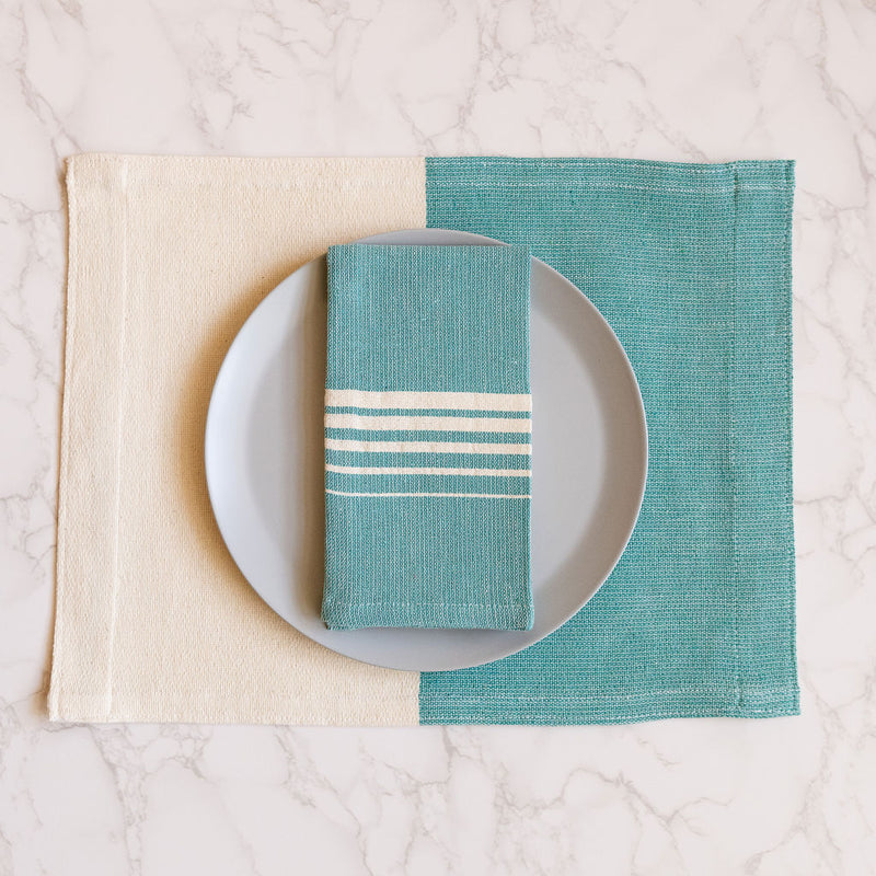 A Teal Casa Oaxaca Cotton Cloth Napkin, featuring five delicate white lines, sits elegantly on a grey plate, perfectly paired with a matching woven placemat. This vibrant, handcrafted napkin adds a touch of artisanal Mexican charm to any eco-friendly table setting.