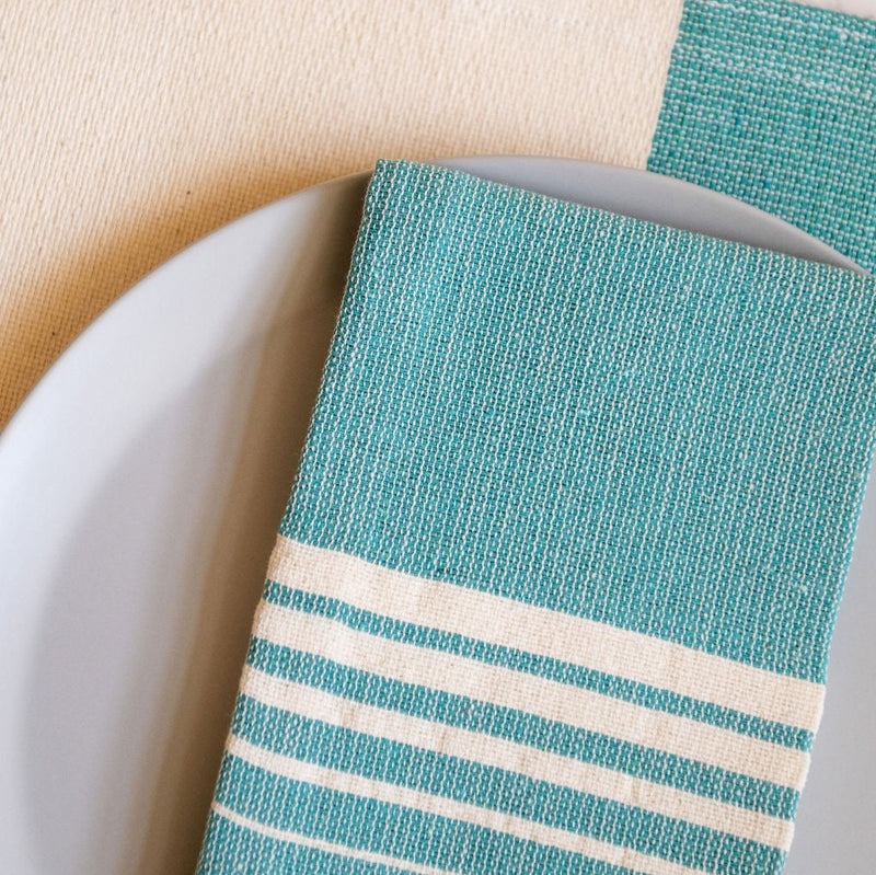 A Teal Casa Oaxaca Cotton Cloth Napkin, featuring five delicate white lines, sits elegantly on a grey plate, perfectly paired with a matching woven placemat. This vibrant, handcrafted napkin adds a touch of artisanal Mexican charm to any eco-friendly table setting.