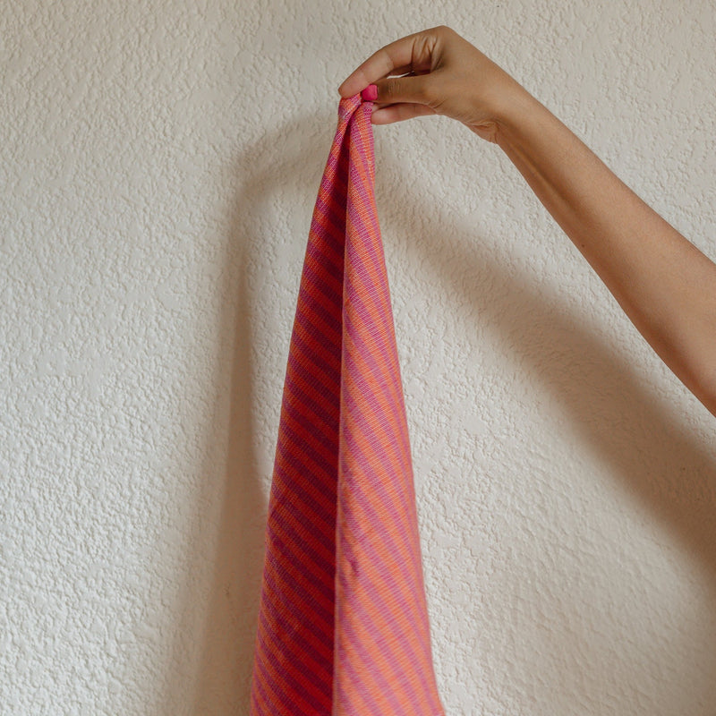 Handwoven cotton napkin in Orange and Pink stripes held by a hand against a wall. Ethically made.