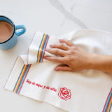 white cleaning cloth embroidered with soy de aqui y de alla wiping down a counter