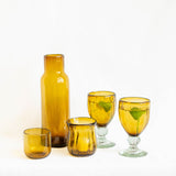 A set of five amber glass tableware pieces, including a tall CARAFE, a small tumbler, a short tumbler, and a fluted handblown glass candle holder, arranged on a white background.