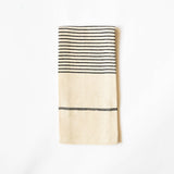 A folded cream-colored linen kitchen towel with black woven stripes, displayed against a clean white background.