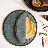 A round fabric hunter green cloth tortilla warmer with a textured green exterior and a stack of corn tortillas inside, shown on a white marble surface with pumpkin seeds, dried red chilies, and a black plate in a styled flat-lay arrangement.