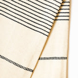 A close-up of a cream-colored woven kitchen hand towel with black dotted and dashed horizontal stripes, folded over on a plain white background.