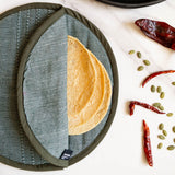 A round hunter green cloth tortilla warmer pouch with a textured gray-green exterior, shown partially open with tortillas inside. The tortilla warmer pouch is photographed from above on a white marble surface with dried red chilies and green pumpkin seeds scattered nearby.