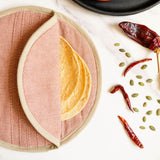 A folded cinnamon brown woven tortilla warmer holding tortillas inside, styled on a white marble surface with dried red chilies, green pumpkin seeds, and seeds on a black plate.