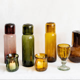 A set of vintage handblown translucent glass candle holders and carafes in amber and olive green, arranged on a white surface against a white background.