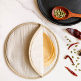 A round beige cloth tortilla warmer pouch with a stitched rim, shown on a white marble surface. A stack of soft corn tortillas is partially folded inside the holder.