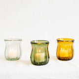 Three ribbed glass candle holders in clear, olive green, and amber yellow, each containing a lit tea light candle, arranged on a white textured fabric background.