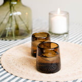 Two chocolate brown glass tumblers with ice and water inside on a woven placemat against a softly blurred background.