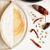 A styled flat-lay of tortillas held warm in a beige cloth tortilla warmer pouch, dried red chiles, and green pumpkin seeds arranged on a white marble surface.