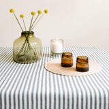 A styled tabletop scene with a hunter green striped tablecloth, a translucent olive-green glass vase holding yellow flower stems, a white candle in a clear holder, and two chocolate brown glass tumblers on a round woven placemat against a plain light beige wall.