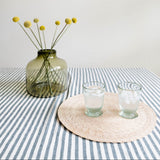 A styled tabletop scene with a translucent olive-green glass vase holding yellow dried flower stems, next to two clear drinking glasses on a round woven placemat over a blue-and-white striped cloth.