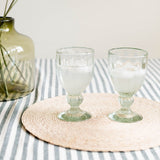 Two clear handblown glass goblets filled with water, placed on a round woven beige placemat over a blue-and-white striped tablecloth.