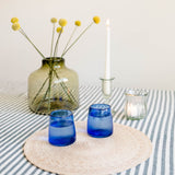 Two cobalt blue glass on a round woven placemat, set on a green and white striped tablecloth next to a green glass vase with yellow flowers, a white candle, and a small glass clear votive holder.